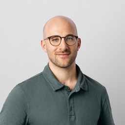 Portrait of a bald man with glasses, smiling at the camera, wearing a navy blue t-shirt, indoors with a blurred background.