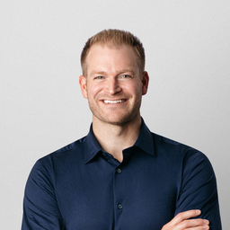 Portrait of a man with short brown hair, smiling at the camera, wearing a black shirt, in an indoor setting with a soft-focus background.