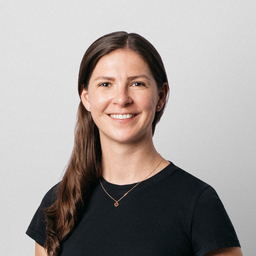 Portrait of a woman with long hair and a friendly smile, wearing a light beige striped sweater, in an indoor environment with a plant in the background.