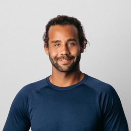 Portrait of a man with curly hair and a beard, smiling gently, wearing a navy blue shirt, with an indoor blurred background featuring greenery.